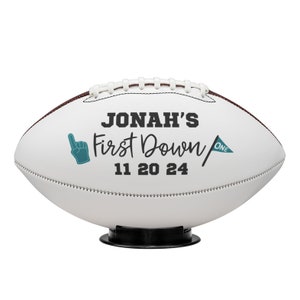 May include: White football with brown stitching and the text "JONAH'S First Down 11 20 24". Includes a teal foam finger and a teal flag with the word "ONE". The football is on a black stand.