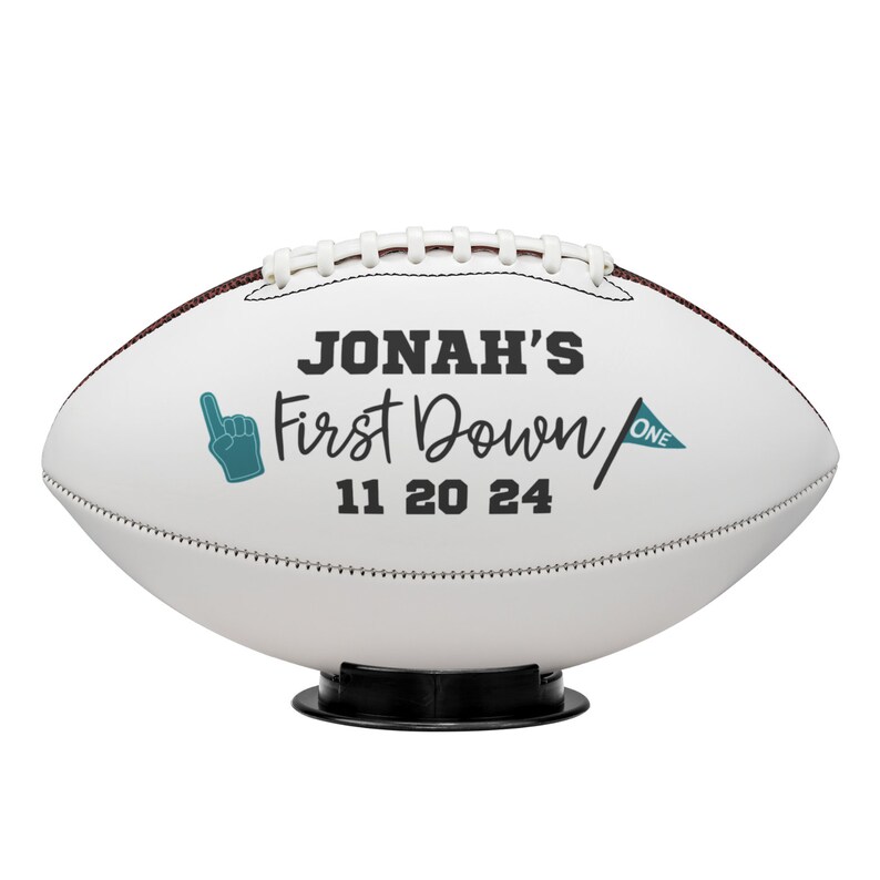 May include: White football with brown stitching and the text "JONAH'S First Down 11 20 24". Includes a teal foam finger and a teal flag with the word "ONE". The football is on a black stand.