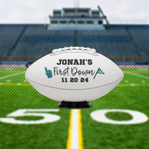 May include: A white football with black text that reads "Jonah's First Down 11 20 24". The football has a teal colored hand pointing up and a teal flag with the word "one". The football is on a black stand.