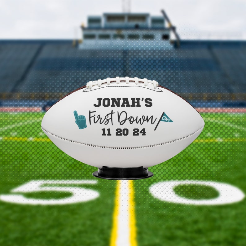 May include: A white football with black text that reads "Jonah's First Down 11 20 24". The football has a teal colored hand pointing up and a teal flag with the word "one". The football is on a black stand.