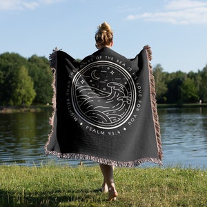 Op de afbeelding: Zwarte plaid met een wit cirkelvormig ontwerp met golven, een maan en sterren. De tekst luidt: 'Machtiger dan de golven van de zee is zijn liefde voor jou' en 'Psalm 93:4'. De deken heeft gefranjerde randen.