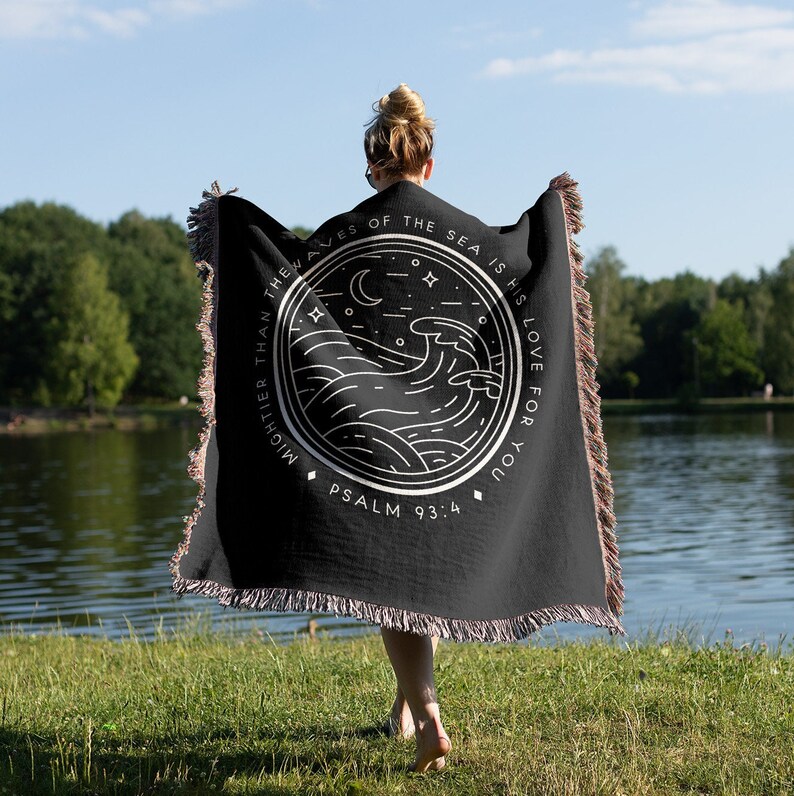 Op de afbeelding: Zwarte plaid met een wit cirkelvormig ontwerp met golven, een maan en sterren. De tekst luidt: 'Machtiger dan de golven van de zee is zijn liefde voor jou' en 'Psalm 93:4'. De deken heeft gefranjerde randen.