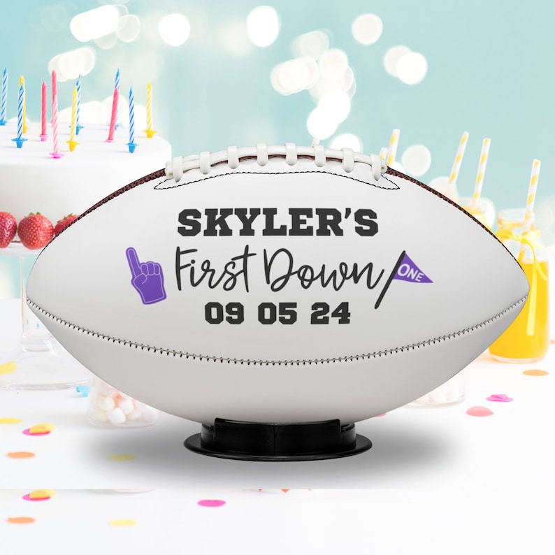 May include: White football with brown stitching, personalized with "Skyler's First Down" and the date 09 05 24. Features a purple foam finger and a purple pennant with "ONE". The football is displayed on a black stand.