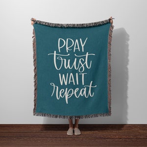 May include: Teal throw blanket with the words "PRAY trust WAIT repeat" in white script. The blanket has a fringed edge in shades of brown and black. The blanket is held up against a white wall and brown wooden floor.