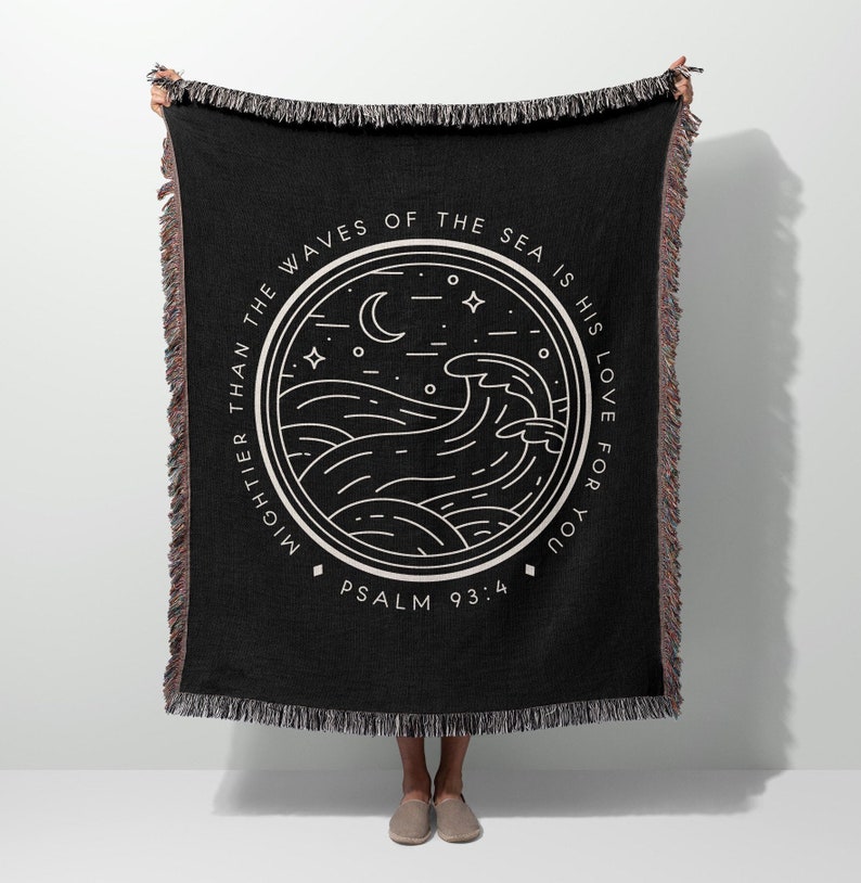 Op de afbeelding: Zwarte geweven sprei met witte tekst en een cirkelvormig ontwerp met golven, een maan en sterren. De tekst luidt "Mightier than the waves of the sea is his love for you" en "Psalm 93:4". De sprei heeft gefranjerde randen.