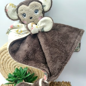 May include: A brown plush monkey security blanket with a white and pink patterned fabric on the back. The monkey has embroidered eyes and a pink nose.