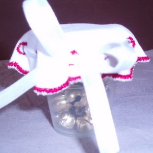May include: A white fabric jar cover with red trim, tied with a white ribbon, and filled with small silver bells.