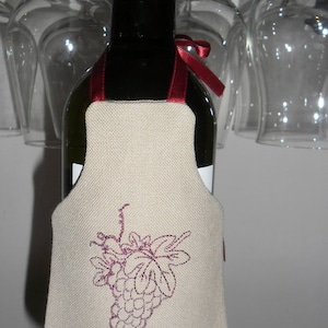 May include: A wine bottle with a beige fabric cover featuring an embroidered design of a bunch of grapes. The cover has a red ribbon tied around the bottle neck.
