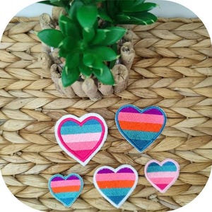 Puede incluir: Cinco parches coloridos en forma de corazón con un diseño de rayas arcoíris. Los parches están hechos de tela y tienen un contorno blanco.