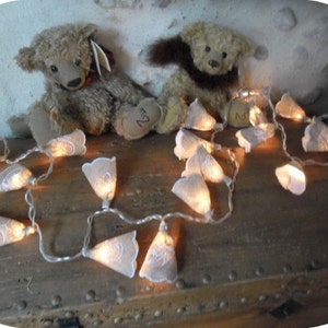 May include: A string of white fabric fairy lights with a warm white glow. The lights are shaped like small bells and are strung on a clear wire. The lights are draped over a wooden surface with two teddy bears in the background.