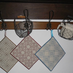 Może przedstawiać: Trzy ozdobne uchwyty do garnków z misternymi wzorami wiszą na haczykach na drewnianej półce. Uchwyty są beżowe z czerwonymi, niebieskimi i brązowymi haftami. Dwa druciane kosze wiszą na półce nad uchwytami.