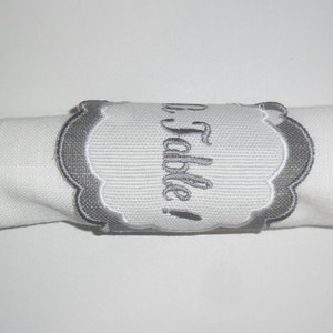 May include: A grey embroidered napkin ring with a white background and the text "A Table!" in a cursive font.