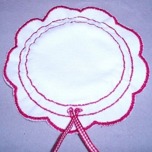 May include: White fabric circle with a scalloped edge and three red stitched lines. The circle has a red and white gingham ribbon tied at the bottom.