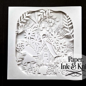 May include: White paper card with a detailed paper cut design of a house in a forest with mushrooms and flowers. The text "Paper, Ink & Knife" is in the bottom right corner.
