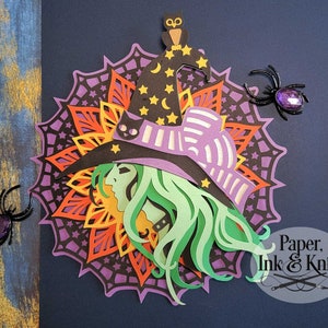 May include: Paper cut-out of a witch with a purple and white hat, green hair, and a black cat on her hat. The witch is surrounded by a mandala with orange, red, and purple colors. The background is dark blue. The image includes the text "Paper, Ink & Knife".