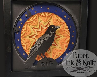 May include: A black raven papercraft sculpture with a blue and orange mandala background. The raven is perched on a tombstone with the word "RIP" inscribed. The artwork is framed in black and has a "Paper, Ink & Knife" logo in the bottom right corner.