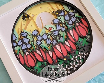 May include: A paper cut art piece in a white frame with a circular design of a spring garden scene. The scene features red and pink tulips, bluebells, green leaves, and bees. The background is a yellow circle with white dots. The text "Paper, Ink & Knife" is at the bottom of the design.