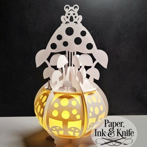 May include: White papercraft lamp featuring a mushroom and frog design. The lamp is illuminated from within, emitting a warm glow. The lamp is crafted from paper and adorned with cut-outs of mushrooms and a frog.