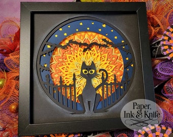 May include: A framed, layered paper art piece featuring a black cat silhouette sitting in front of a fence, with a full moon and bats in the background. The moon is a layered mandala design in shades of orange and yellow, set against a dark blue sky with stars.