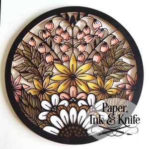 May include: A circular wall art piece with a black frame and a paper cut design of flowers in shades of pink, yellow, and white. The design features a variety of flowers, including daisies, lilies, and roses. The text "Paper, Ink & Knife" is visible in the lower right corner of the image.