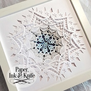 May include: A white framed paper art piece with a detailed snowflake design in shades of blue and white. The artwork is titled "Paper, Ink & Knife".