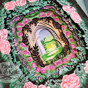 May include: A layered paper art piece featuring a garden scene with a stone archway. The artwork is framed by pink roses and green foliage, with the text "Paper, Ink & Knife" in the lower left corner.