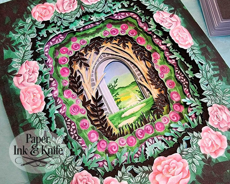 May include: A layered paper art piece featuring a garden scene with a stone archway. The artwork is framed by pink roses and green foliage, with the text "Paper, Ink & Knife" in the lower left corner.