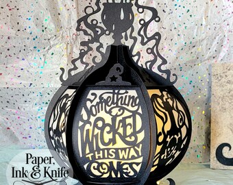 May include: Black paper lantern shaped like a cauldron with the words "Something Wicked This Way Comes" cut out. The lantern is lit from inside, casting a warm glow.