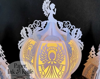 May include: A white paper lantern with an angel design, illuminated from within. The lantern features intricate cut-out patterns and is topped with a silhouette of an angel. The text "Paper, Ink & Knife" is visible on the side.