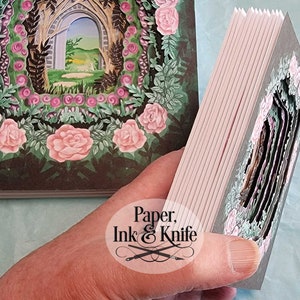 May include: A handmade paper art book with a layered design. The cover and interior pages feature a garden scene with pink roses, green foliage, and an arched doorway. The book is held open, revealing the intricate cut-out layers.
