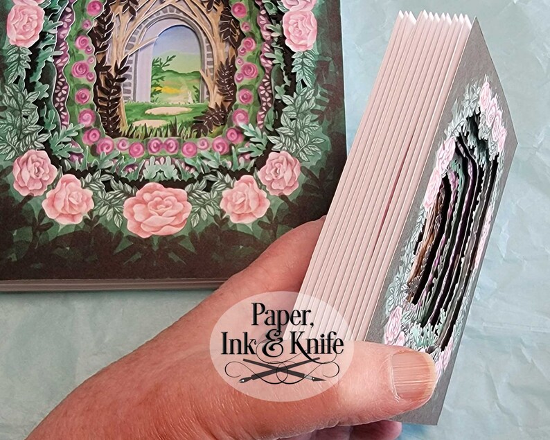 May include: A handmade paper art book with a layered design. The cover and interior pages feature a garden scene with pink roses, green foliage, and an arched doorway. The book is held open, revealing the intricate cut-out layers.