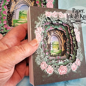May include: A stack of paper art cards featuring a layered garden scene with a stone archway, surrounded by pink roses and green foliage. The cards have a dark gray border and the text "Paper, Ink & Knife".