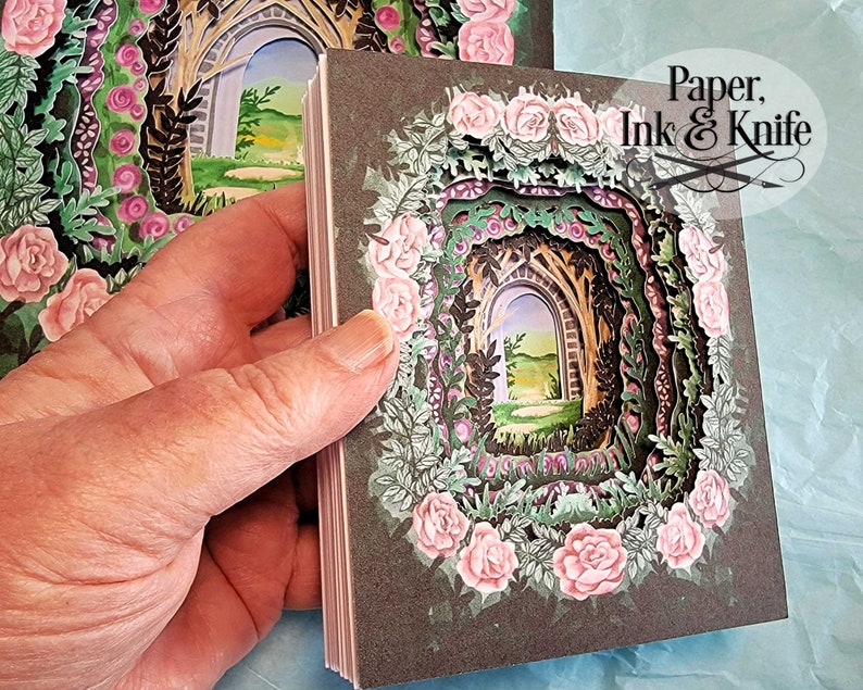 May include: A stack of paper art cards featuring a layered garden scene with a stone archway, surrounded by pink roses and green foliage. The cards have a dark gray border and the text "Paper, Ink & Knife".