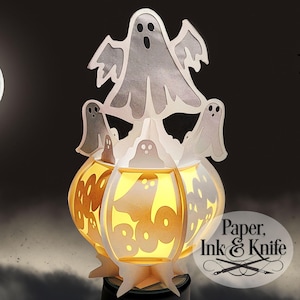 May include: A white papercraft Halloween lantern shaped like a cauldron with ghosts and the word "BOO" on it. The lantern is lit from inside, casting a warm glow.