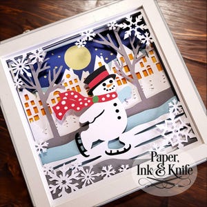 May include: A white snowman wearing a red and white polka dot scarf and a black top hat is ice skating on a blue and white ice rink. The scene is set in a winter wonderland with white snowflakes and trees. The background is a dark blue sky with a yellow moon and white stars. The text "Paper, Ink & Knife" is in the bottom right corner of the image.