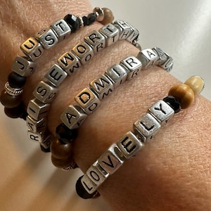 Könnte beinhalten: Vier Armbänder mit Holzperlen und silbernen Buchstabenperlen, die die Wörter "JUST", "RAISE", "WORTH", "ADMIR" und "LOVELY" buchstabieren.