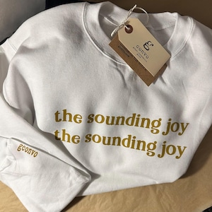 May include: White sweatshirt with the text "the sounding joy" in gold lettering. The brand name "gconvo" is printed on the sleeve. A brown tag is attached to the collar.