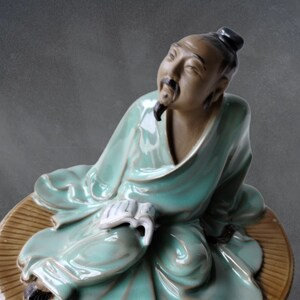 May include: A ceramic figurine of a man sitting cross-legged, wearing a teal robe and holding a book. He has a long beard and a contemplative expression. The figurine is sitting on a brown circular base.