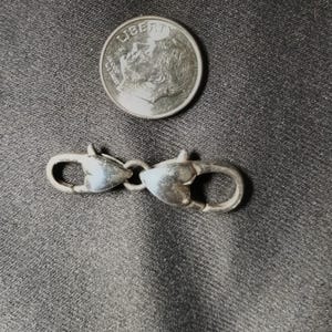May include: A silver-colored clasp with two heart-shaped ends connected by a small ring. A dime is next to the clasp for size comparison. The clasp is likely for jewelry or other small accessories.