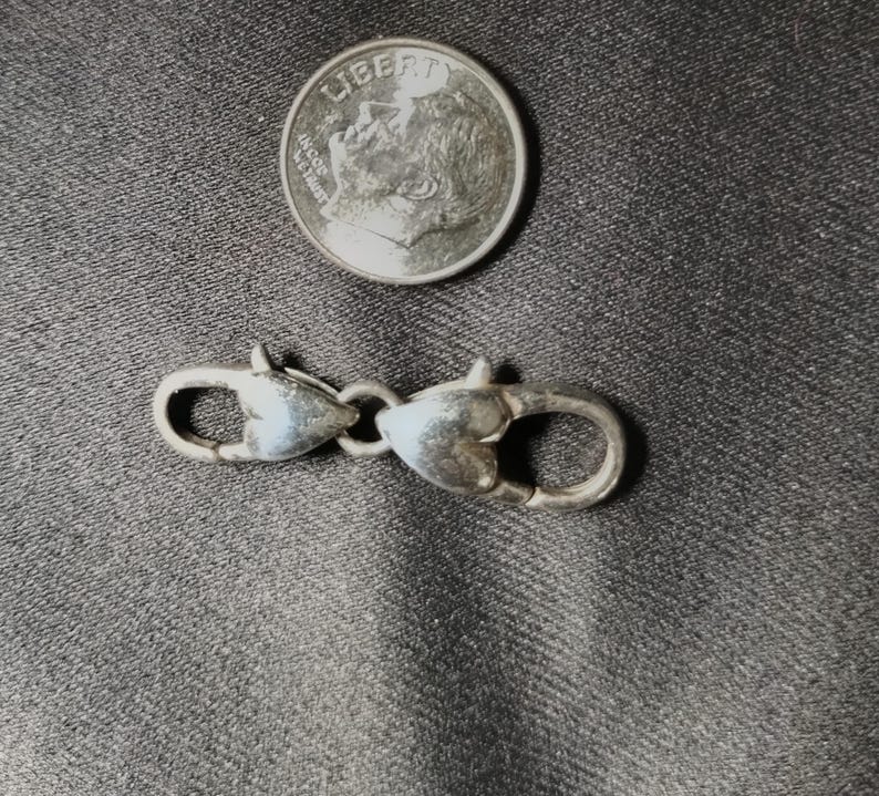 May include: A silver-colored clasp with two heart-shaped ends connected by a small ring. A dime is next to the clasp for size comparison. The clasp is likely for jewelry or other small accessories.