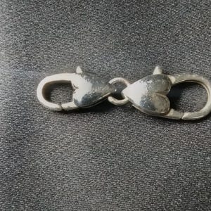 May include: A silver-colored clasp with heart-shaped details. The clasp is designed to connect two parts, featuring a spring-loaded mechanism for secure closure. The metal has a polished finish, reflecting light.