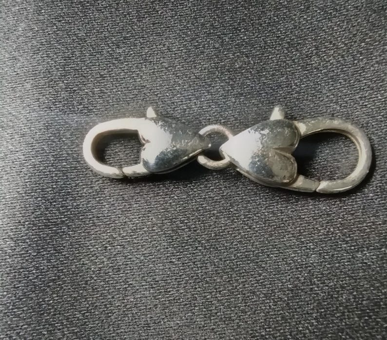 May include: A silver-colored clasp with heart-shaped details. The clasp is designed to connect two parts, featuring a spring-loaded mechanism for secure closure. The metal has a polished finish, reflecting light.