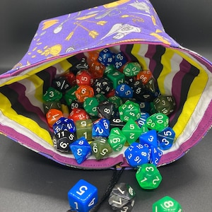May include: A purple fabric dice bag with a yellow, black, and white striped lining. The bag is filled with a variety of colorful dice, including green, blue, red, and black. The bag is tied with a black drawstring and has a skull-shaped charm.