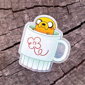 May include: A sticker featuring Jake the Dog from Adventure Time inside a white mug. The mug has a red flower design and a mint green base. Jake is yellow with black eyes and a small smile.