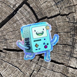 May include: A holographic sticker featuring the character BMO from Adventure Time. The light blue, rectangular gaming console character has a smiling face, thin limbs, and a small yellow star above its head. The sticker has a shimmering finish.