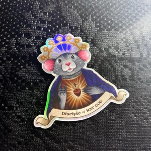 May include: A sticker featuring a cartoon rat with a halo and a heart-shaped emblem. The rat is wearing a blue cape and the text "Disciple of Rat God" is on a banner below.