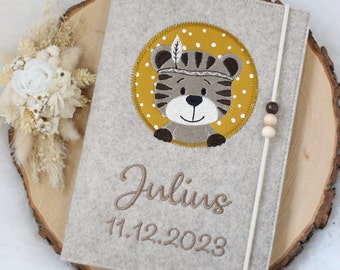 May include: A light gray felt notebook cover featuring an embroidered circular patch with a brown tiger face on a mustard yellow background with white dots. The name Julius and the date 11.12.2023 are embroidered below. A white elastic band with three beads.