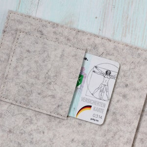 Könnte beinhalten: Ein graues Filzportemonnaie mit einem Fach für eine Karte. Die Karte ist weiß mit einer Zeichnung eines Mannes in einem Kreis und dem Text "VERSICHERTEN KARTE 03.14 gültig bis"