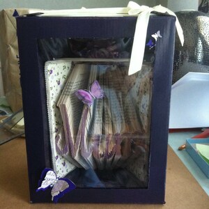 May include: A purple book art sculpture of the word "Mary" in a clear glass display box with a white ribbon and purple butterflies.