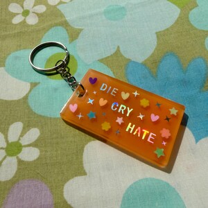 May include: A rectangular orange keychain featuring the holographic text DIE CRY HATE. The surface is decorated with small colorful hearts, stars, and flower shapes. A silver metal split ring is attached to the top corner of the orange rectangle.
